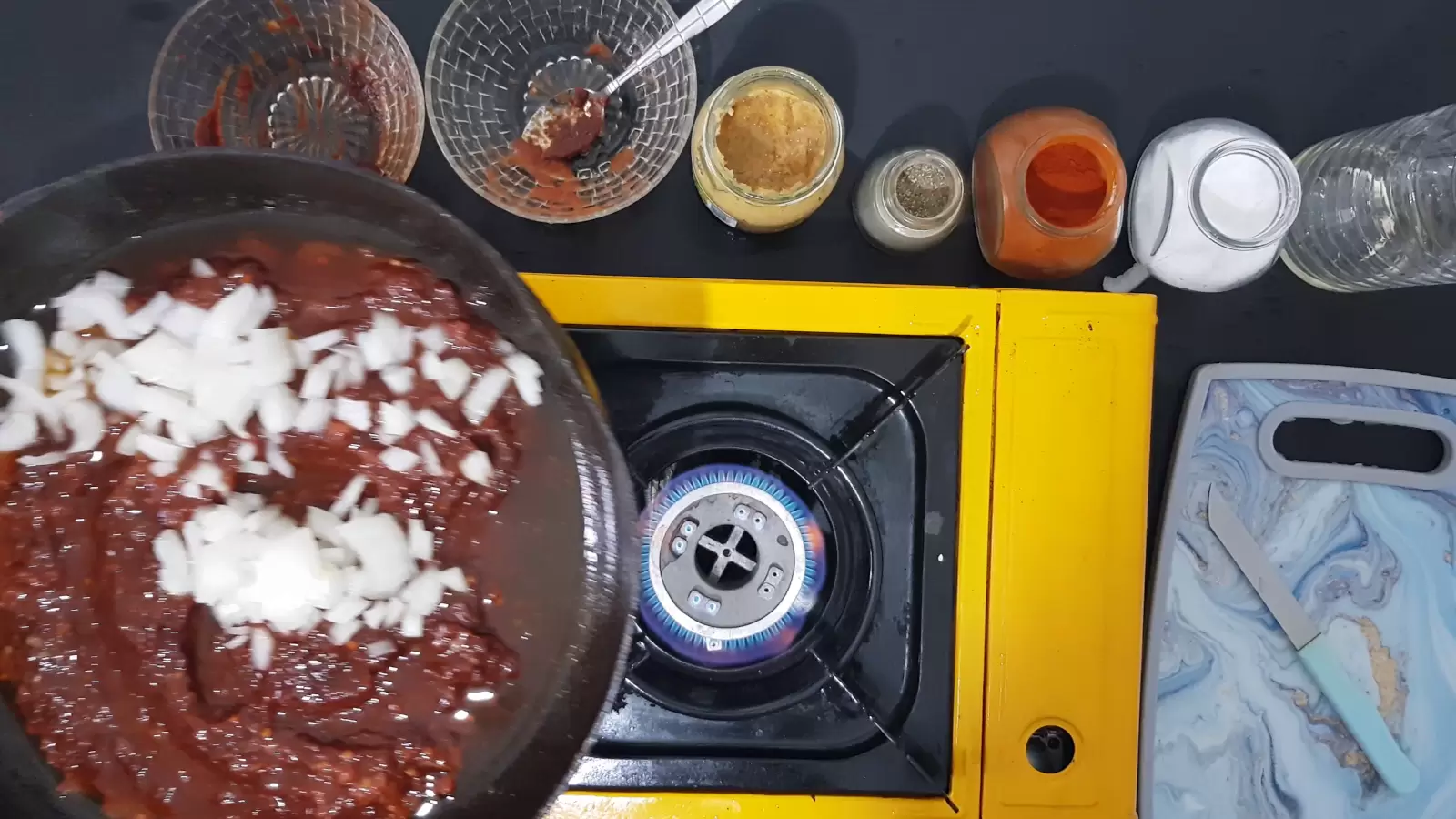 A jar of salt, a jar of red paprika, a jar of black pepper, a jar of crushed garlic, 2 empty bowls, a pan with tomato sauce and chopped onions, a bottle of oil, a cutting board, a lit fire