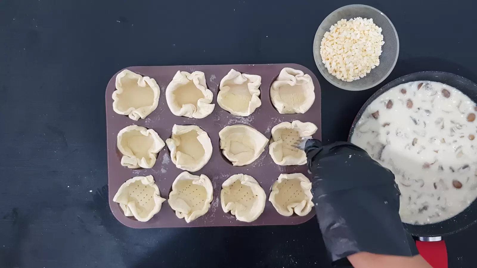 Pan with mushroom and cream filling, bowl with grated cheese, muffin tin with puff pastry squares and fork
