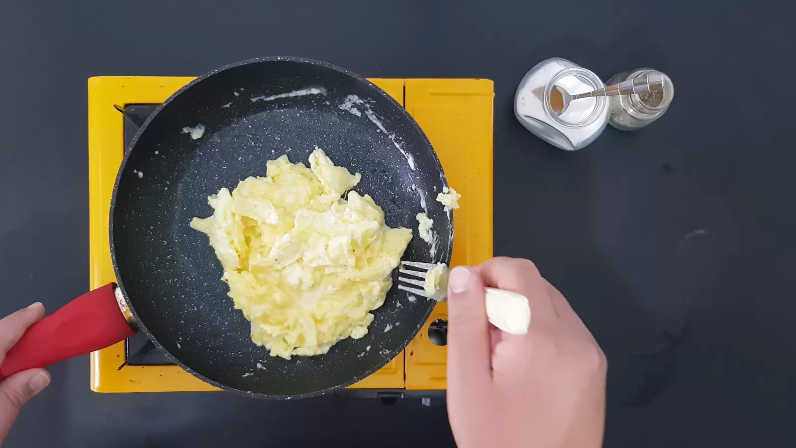 Omelette in a pan, a jar of salt with a spoon, a jar of pepper