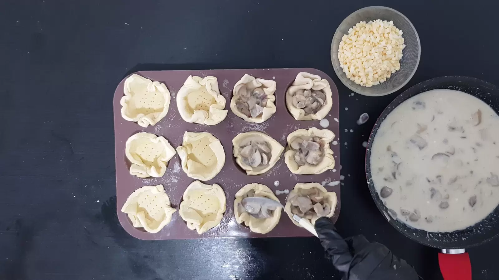 Pan with mushroom and cream filling, bowl with grated cheese, muffin tin with puff pastry squares and filling