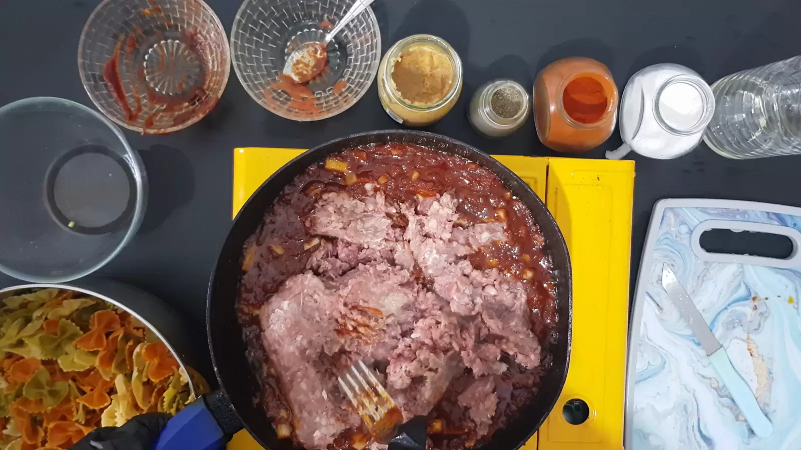 A jar of salt, a jar of red paprika, a jar of black pepper, a jar of crushed garlic, 3 empty bowls, a bowl with farfalle pasta, a pan with tomato sauce and ground meat, a fork, a bottle of oil, a cutting board