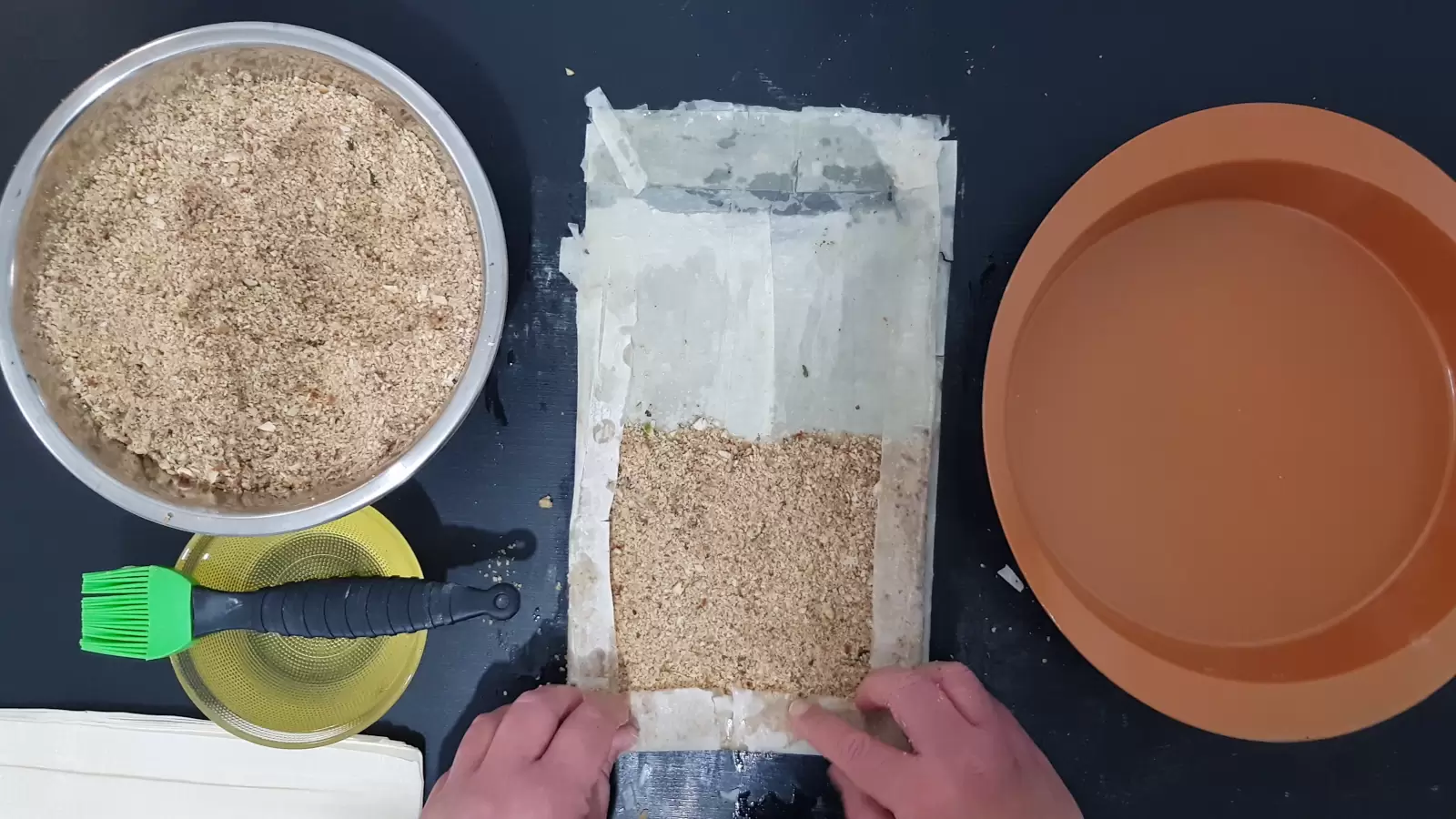 An oven tray, sheets of phyllo with the nut mixture on it, folded on the sides, a bowl with oil and a brush for spreading, a bowl with a mixture of chopped nuts