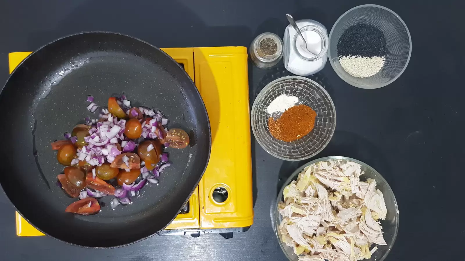 Spices, chicken sliced into strips, sesame, tomatoes and red onion in a pan