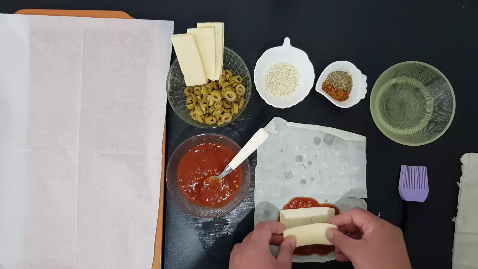 Bowl with oil, bowl with spices, bowl with sesame seeds, bowl with olives and yellow cheese, bowl with tomato sauce, filo dough with tomato sauce and cheese, brush, spoon and baking sheet with baking paper