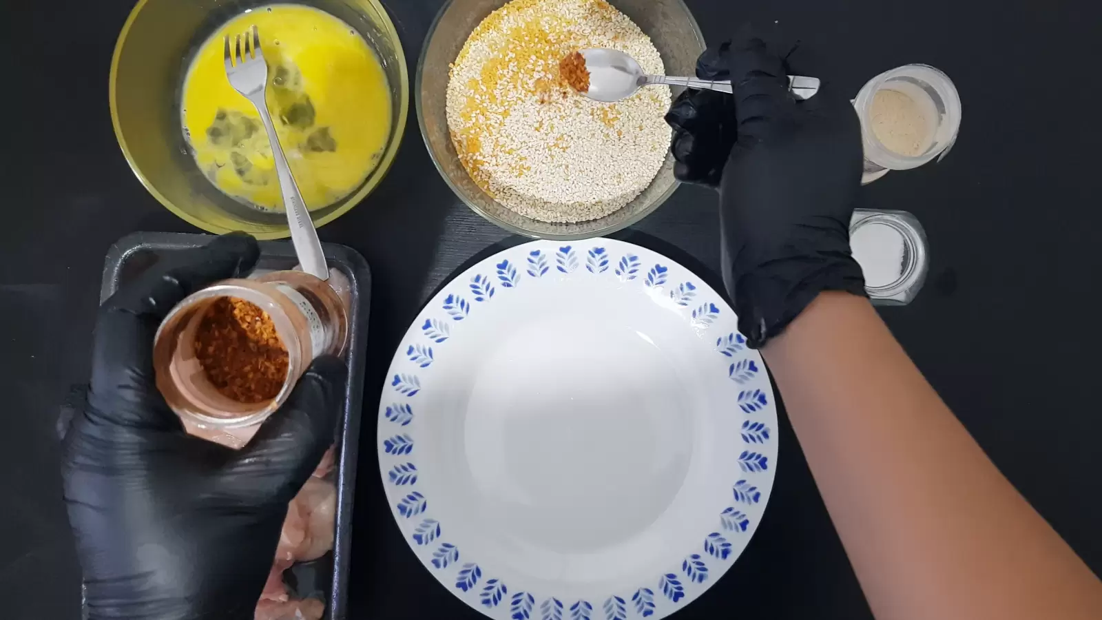 A bowl with eggs and a fork, a bowl with sesame seeds and breadcrumbs, a jar of chili and a spoon, a jar of garlic powder, a jar of salt, a jar of pepper, a plate