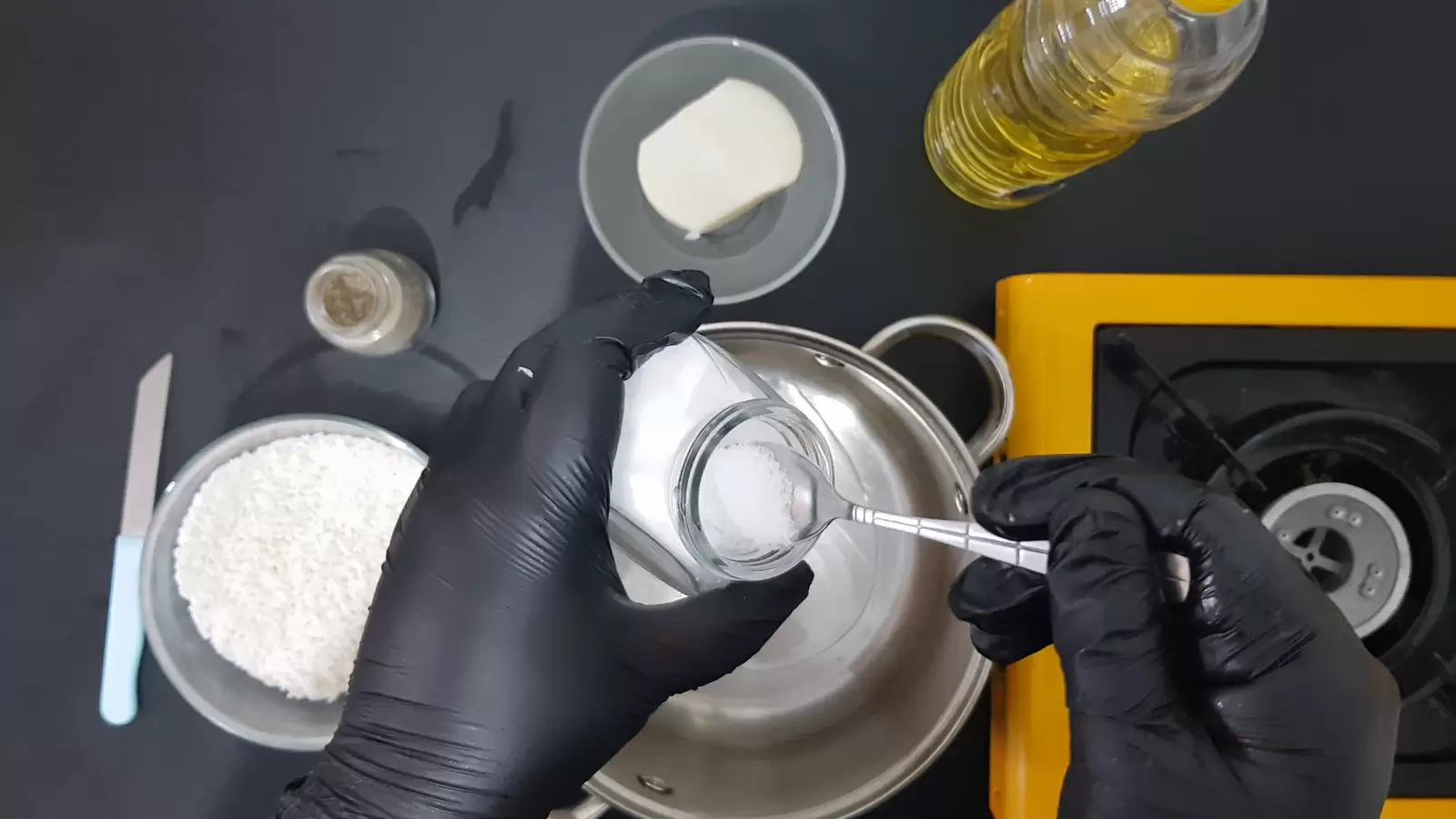 bottle of oil, bowl with onion, jar of salt with spoon, jar of black pepper, bowl with rice, knife, pot