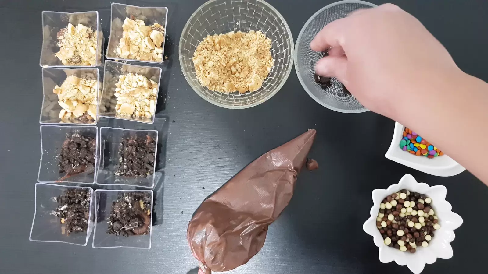 Dessert cups with cookie crumbs, bowl with biscuit crumbs, piping bag with pudding, bowl with candies