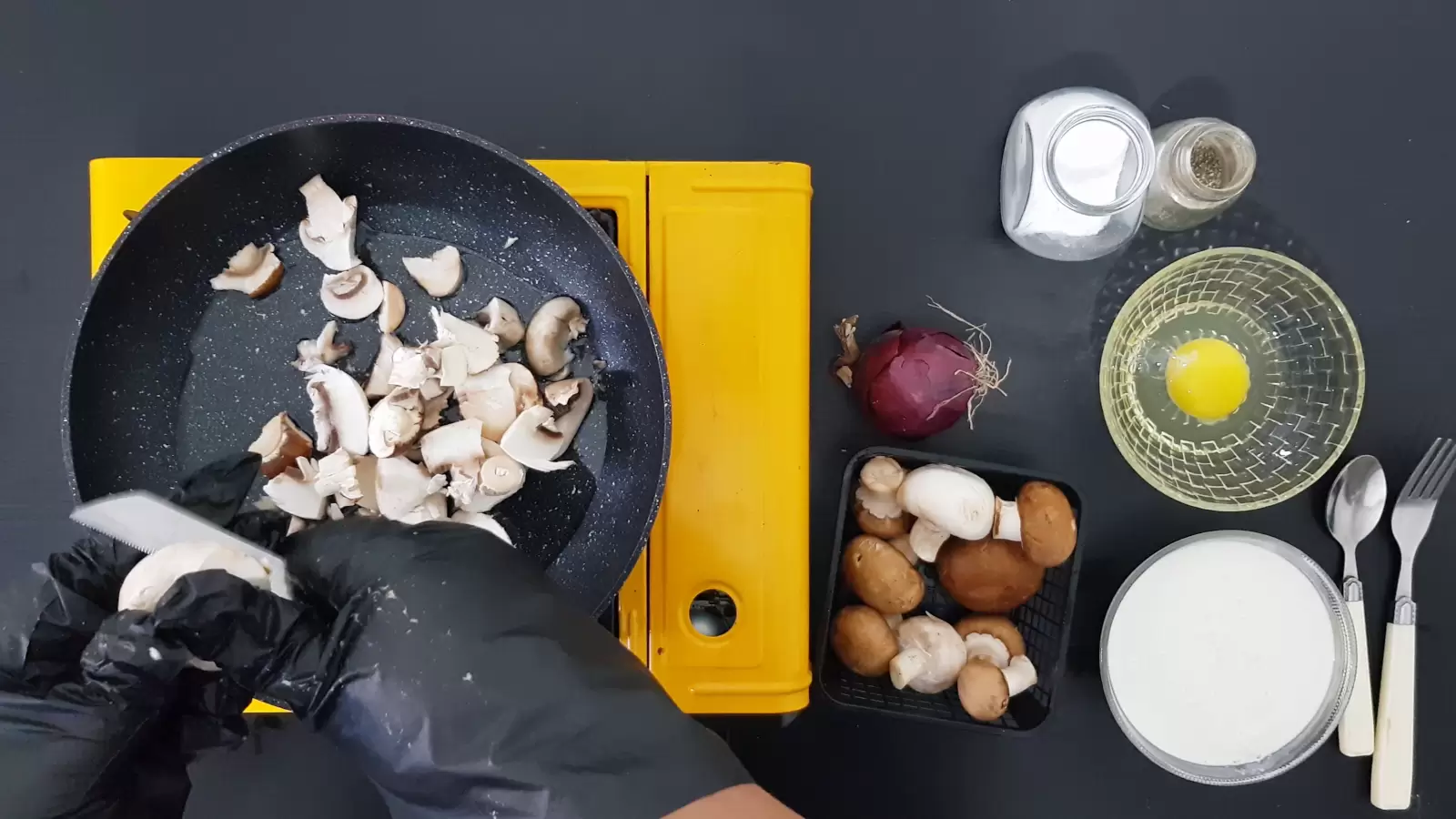 Salt jar, pepper jar, bowl with egg, bowl with cooking cream, red onion, pan with sliced mushrooms, spoon, teaspoon, knife