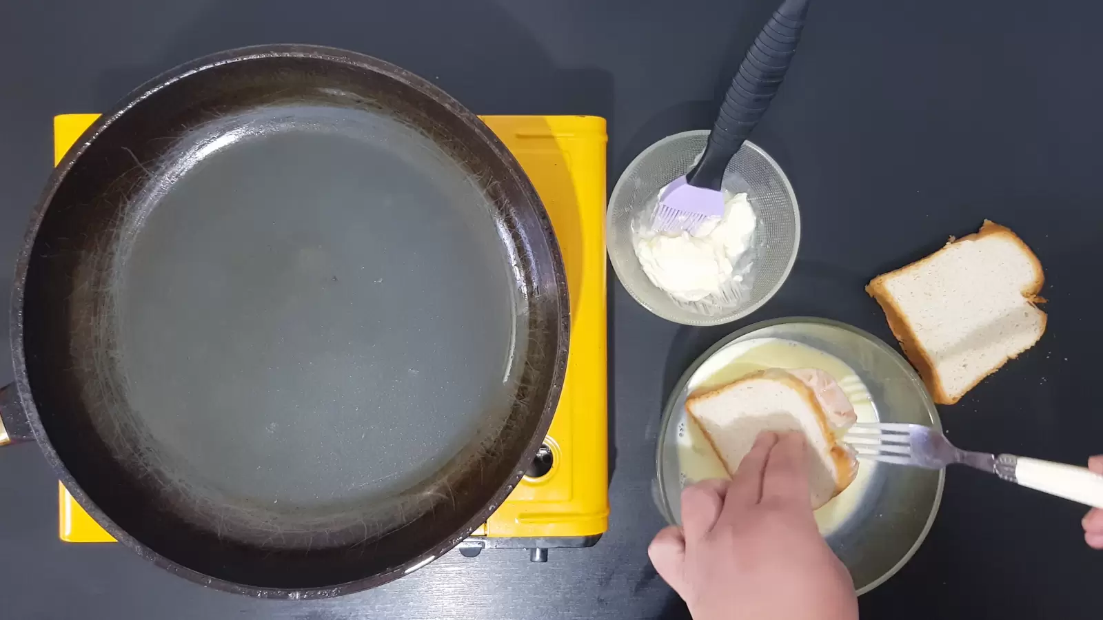 Slice of bread, bowl with butter and brush for brushing, bowl with batter and dipped slice of bread, fork, frying pan