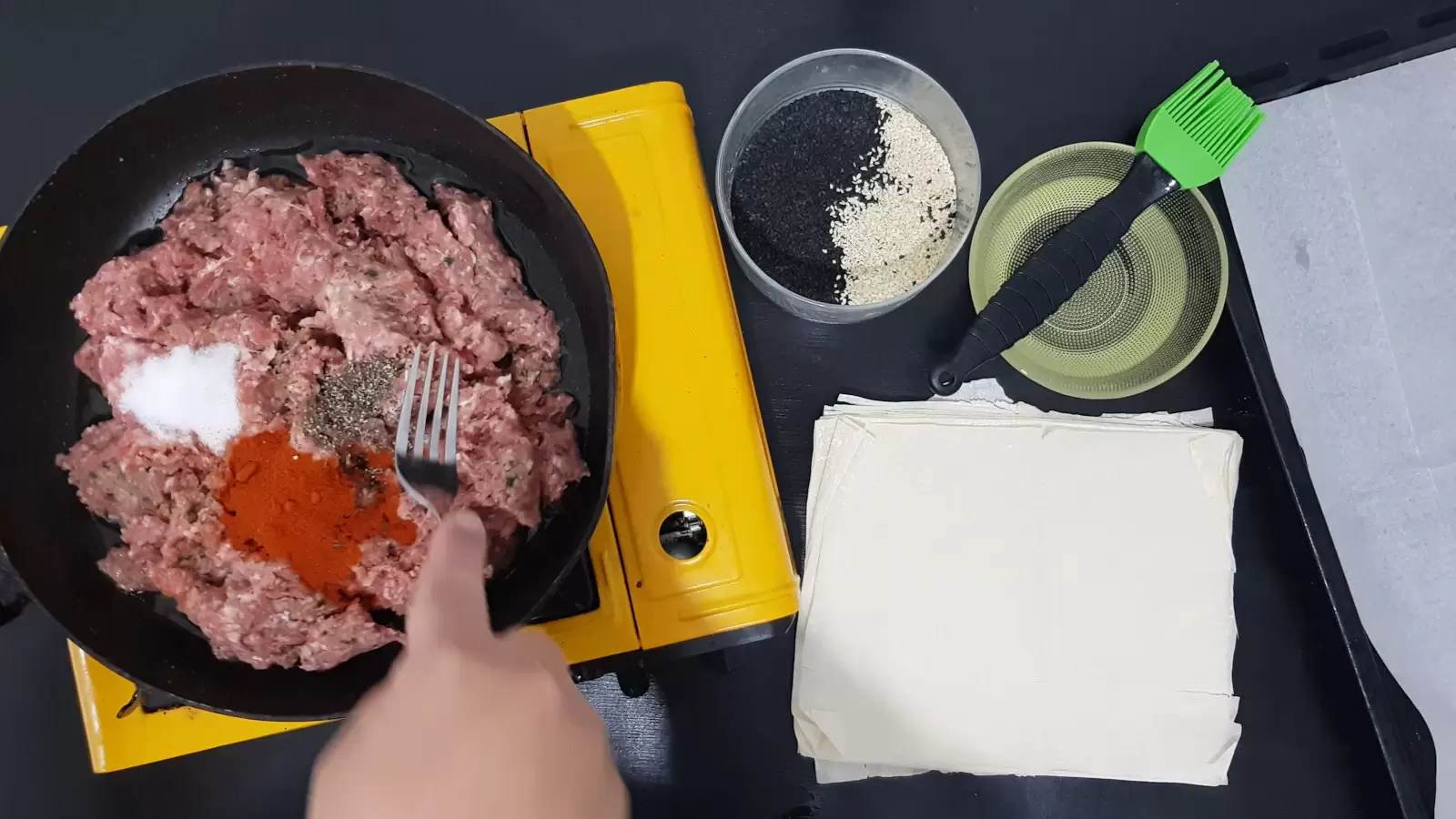 Pan with beef and spices, fork, bowl with oil and a brush for spreading, bowl with white and black sesame seeds, phyllo sheets