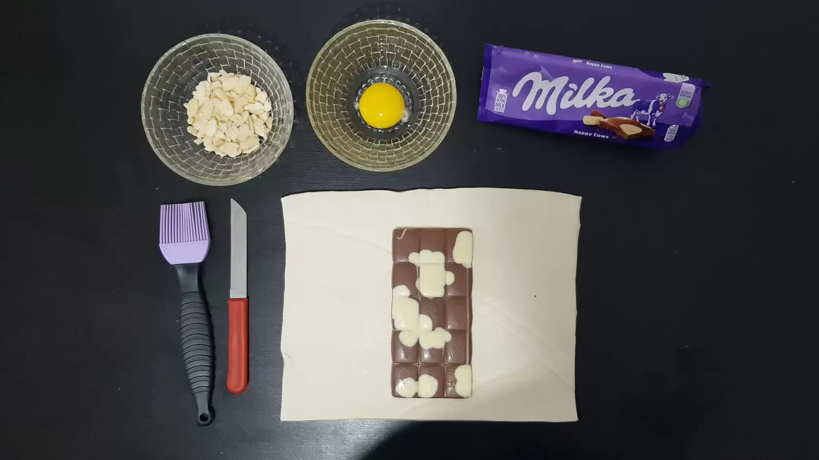 pack of Milka chocolate, bowl with egg, bowl with almonds, puff pastry with Milka chocolate, knife, brush for spreading