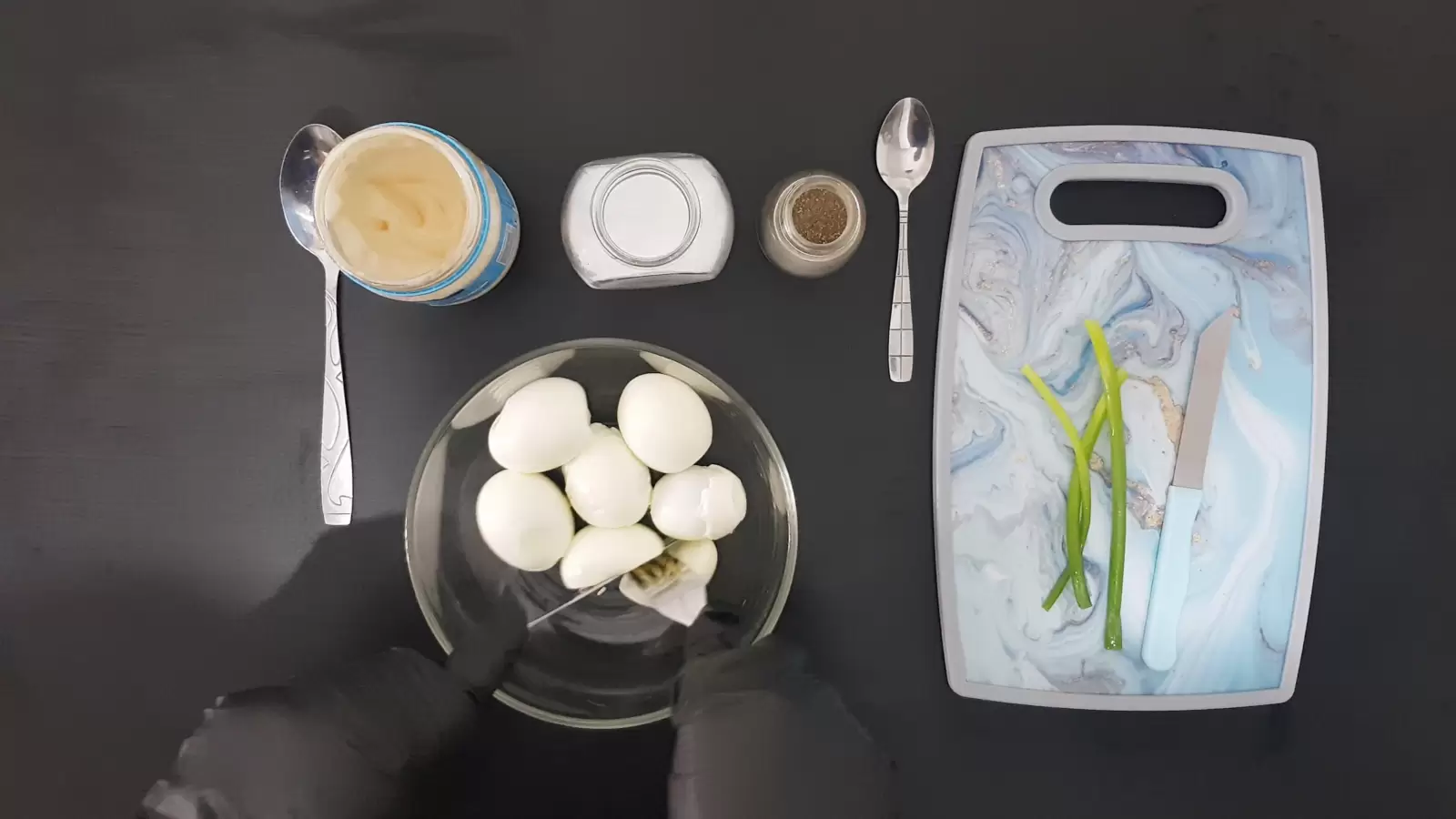 Cutting board with green onions and knife, spoon, black pepper jar, salt jar, mayonnaise jar and spoon, bowl with eggs and knife and fork