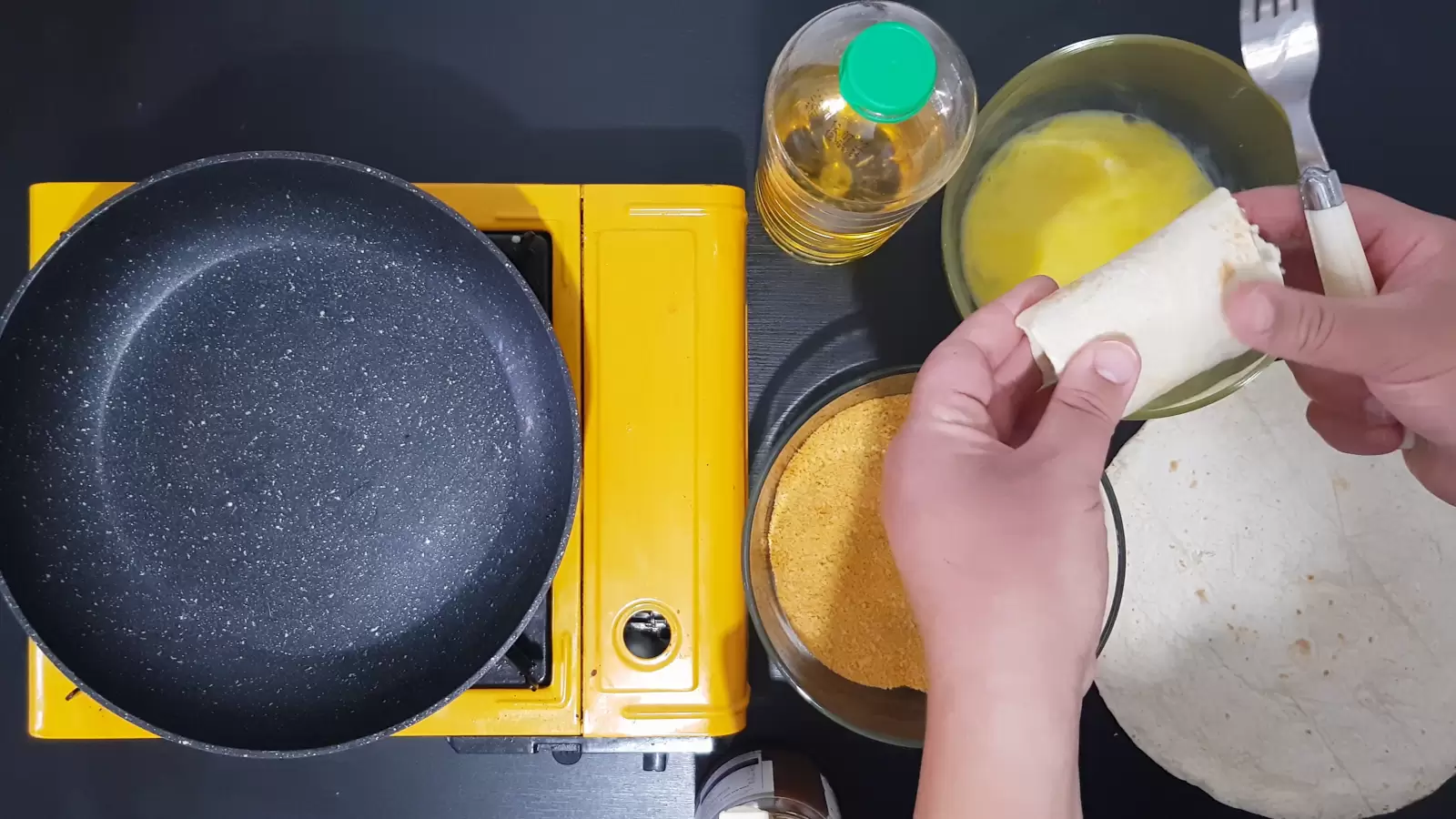Bowl with egg, bottle of oil, bowl with breadcrumbs, tortilla filled with chocolate, stove and frying pan