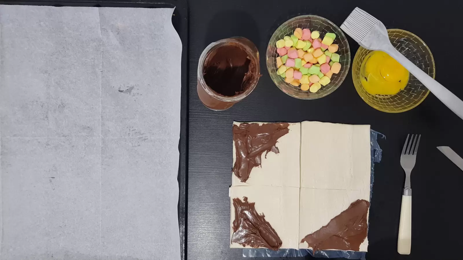 A bowl with an egg and a brush for spreading, a bowl with marshmallows, chocolate spread, a baking sheet with baking paper, puff pastry divided into squares with chocolate spread on each end, a knife and a fork