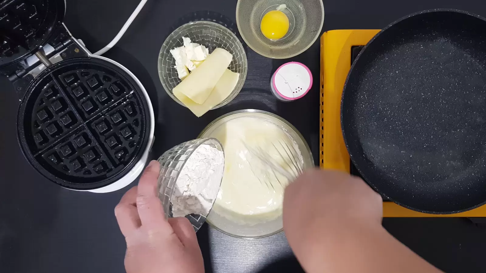 Whisk in a bowl, flour is poured into a bowl, a bowl with an egg, a bowl with cheeses. pan. Waffle maker.