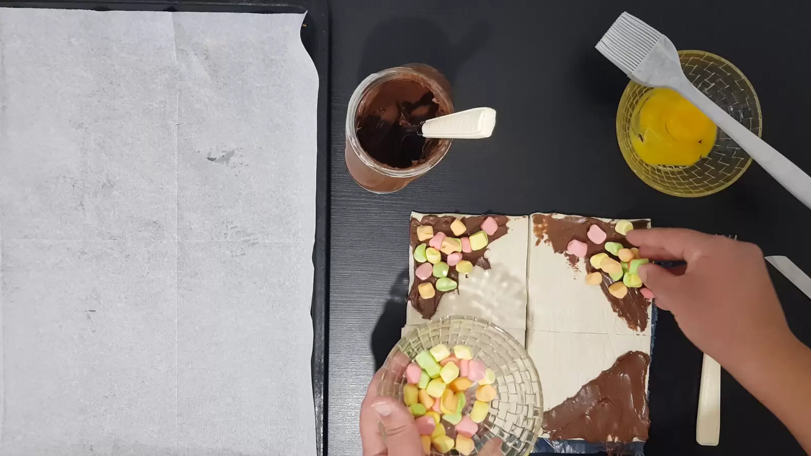 A bowl with an egg and a brush for spreading, a bowl with marshmallows, chocolate spread, a baking sheet with baking paper, puff pastry divided into squares with chocolate and marshmallow spread on each end, a knife and a fork