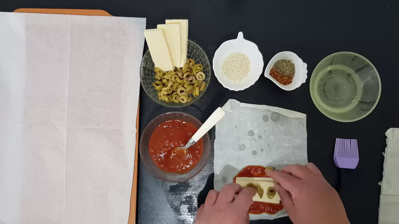 Bowl with oil, bowl with spices, bowl with sesame seeds, bowl with olives and yellow cheese, bowl with tomato sauce, filo dough with tomato sauce cheese and olives, brush, spoon and baking sheet with baking paper