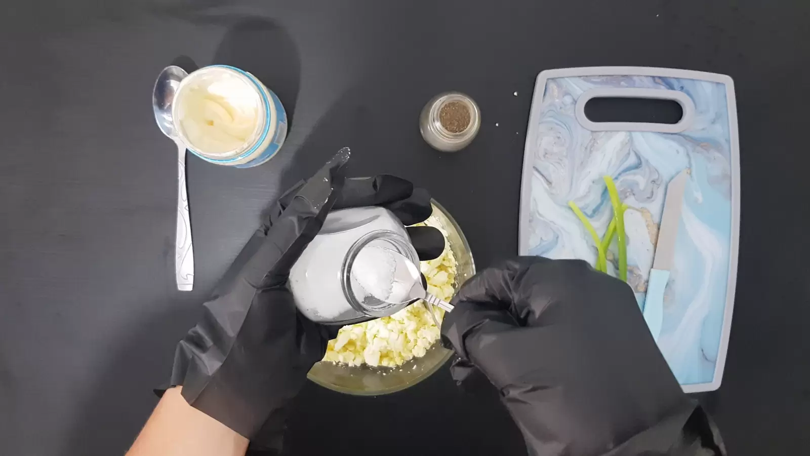 Cutting board with green onions and knife, spoon, black pepper jar, jar and spoon in salt jar, mayonnaise jar and spoon, bowl with eggs