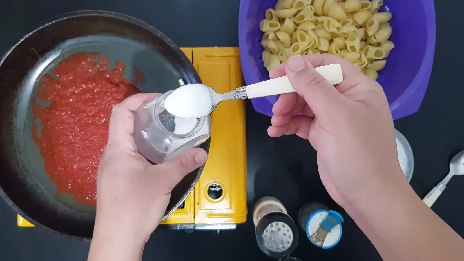 A pan with crushed tomatoes, a bowl with pasta, a teaspoon of salt, a jar of minced garlic, a jar of black pepper, a spoon