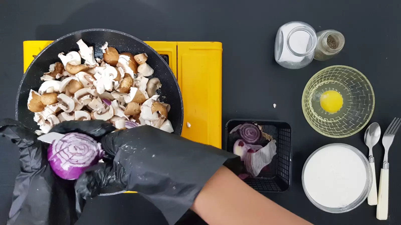 Salt jar, pepper jar, bowl with egg, bowl with cooking cream, red onion, pan with mushrooms, fork, spoon, knife