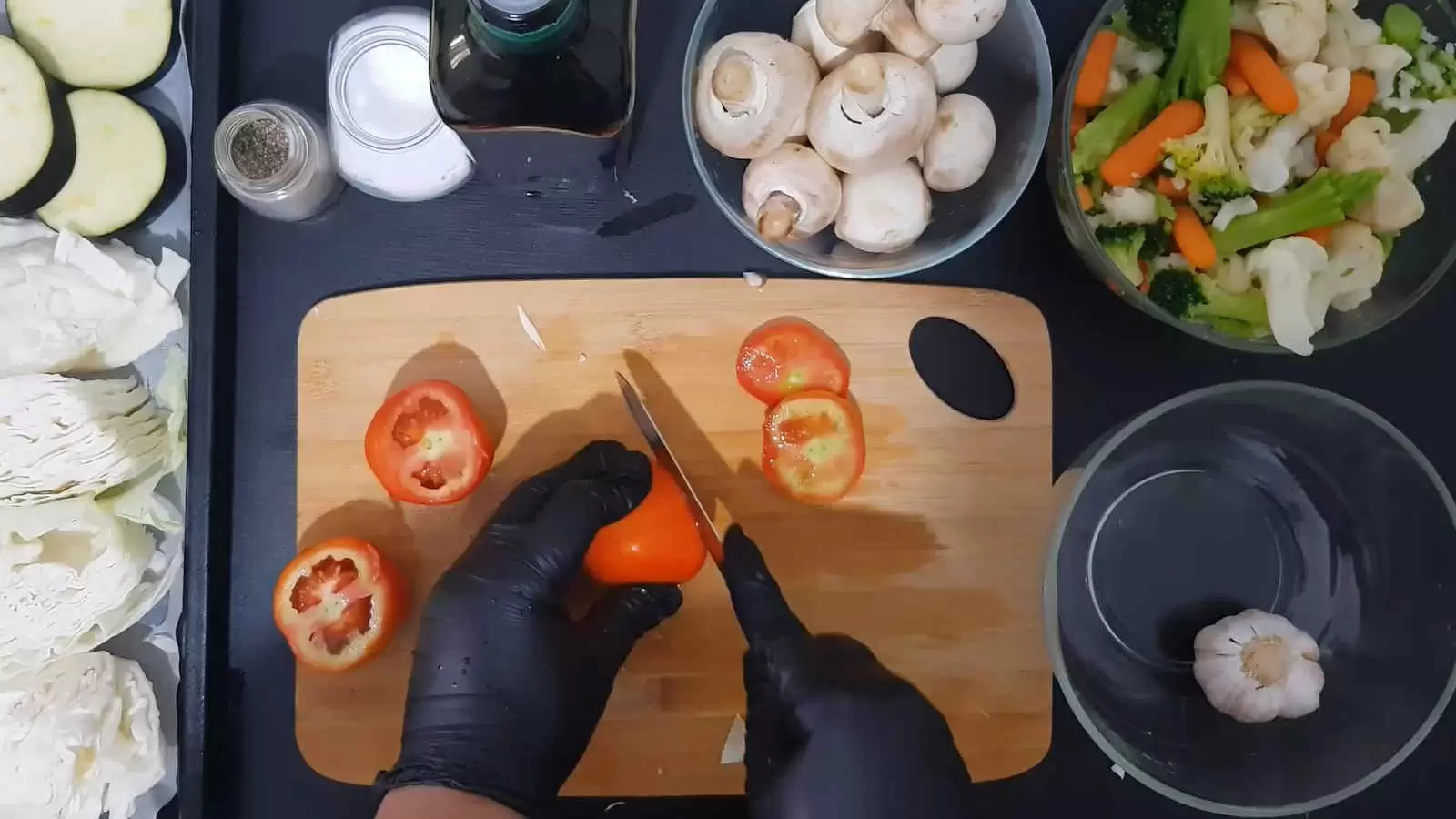 A bowl with cauliflower, carrots and broccoli, a bowl with mushrooms, a bottle of olive oil, a jar of salt, a jar of pepper, a bowl with garlic. Cutting board with tomatoes and knife