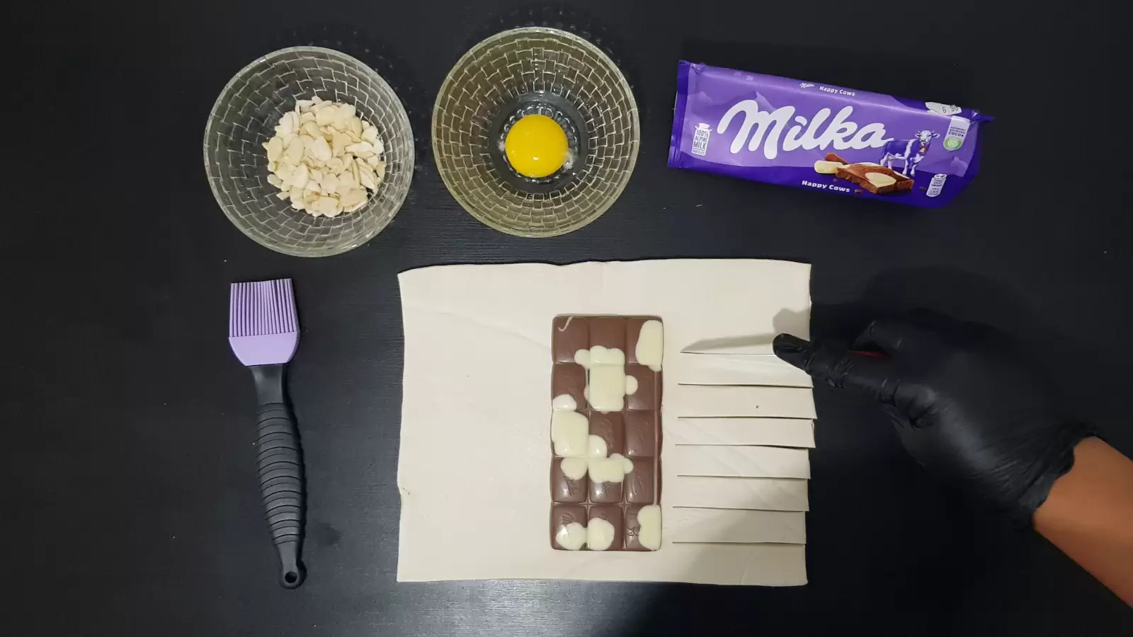 pack of Milka chocolate, bowl with egg, bowl with almonds, puff pastry with Milka chocolate, knife that cuts into strips on the right side, brush for spreading