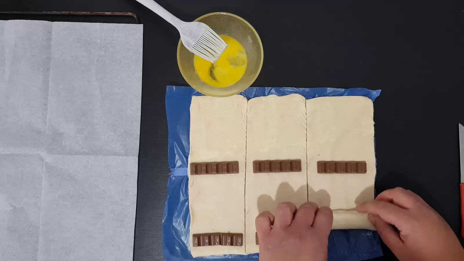 Six Kinder Bueno, rolled puff pastry, a bowl with an egg and a brush, baking paper in an oven tray