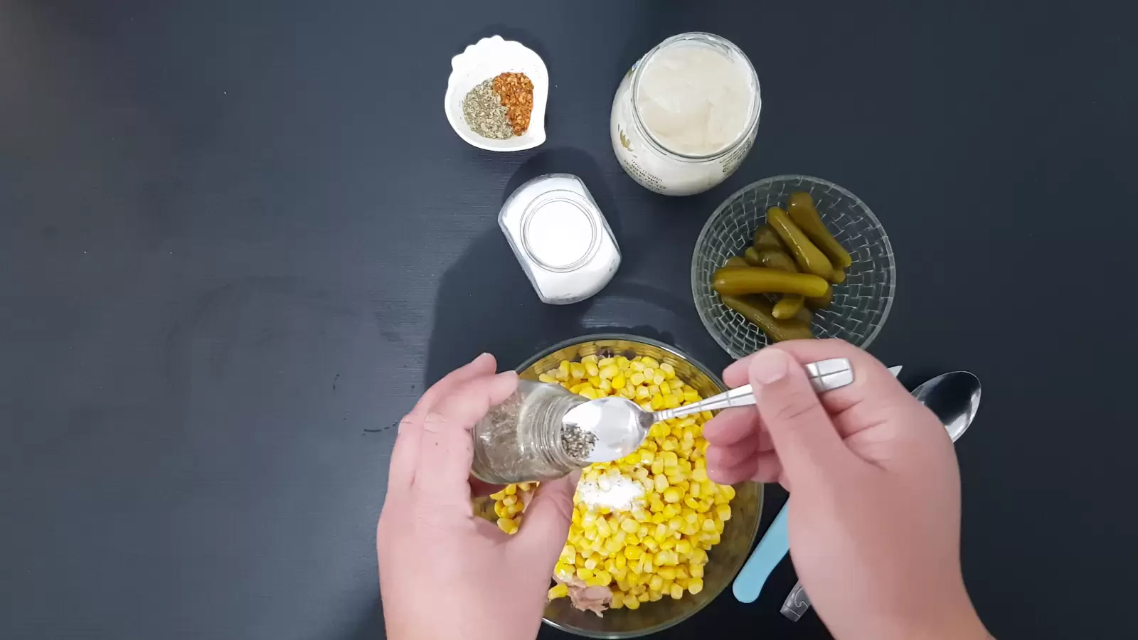 A jar of mayonnaise, a bowl with chili, a jar of salt, a jar of black pepper and a spoon, a bowl with corn, a spoon