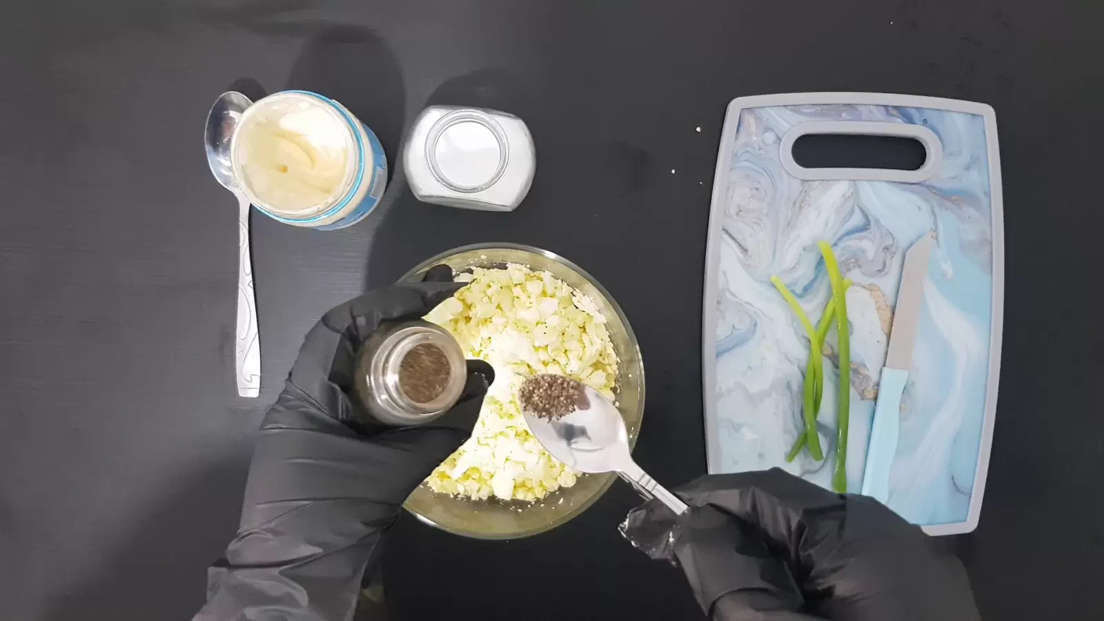 Cutting board with green onions and knife, spoon, salt jar, jar and spoon in black pepper jar, mayonnaise jar and spoon, bowl with eggs