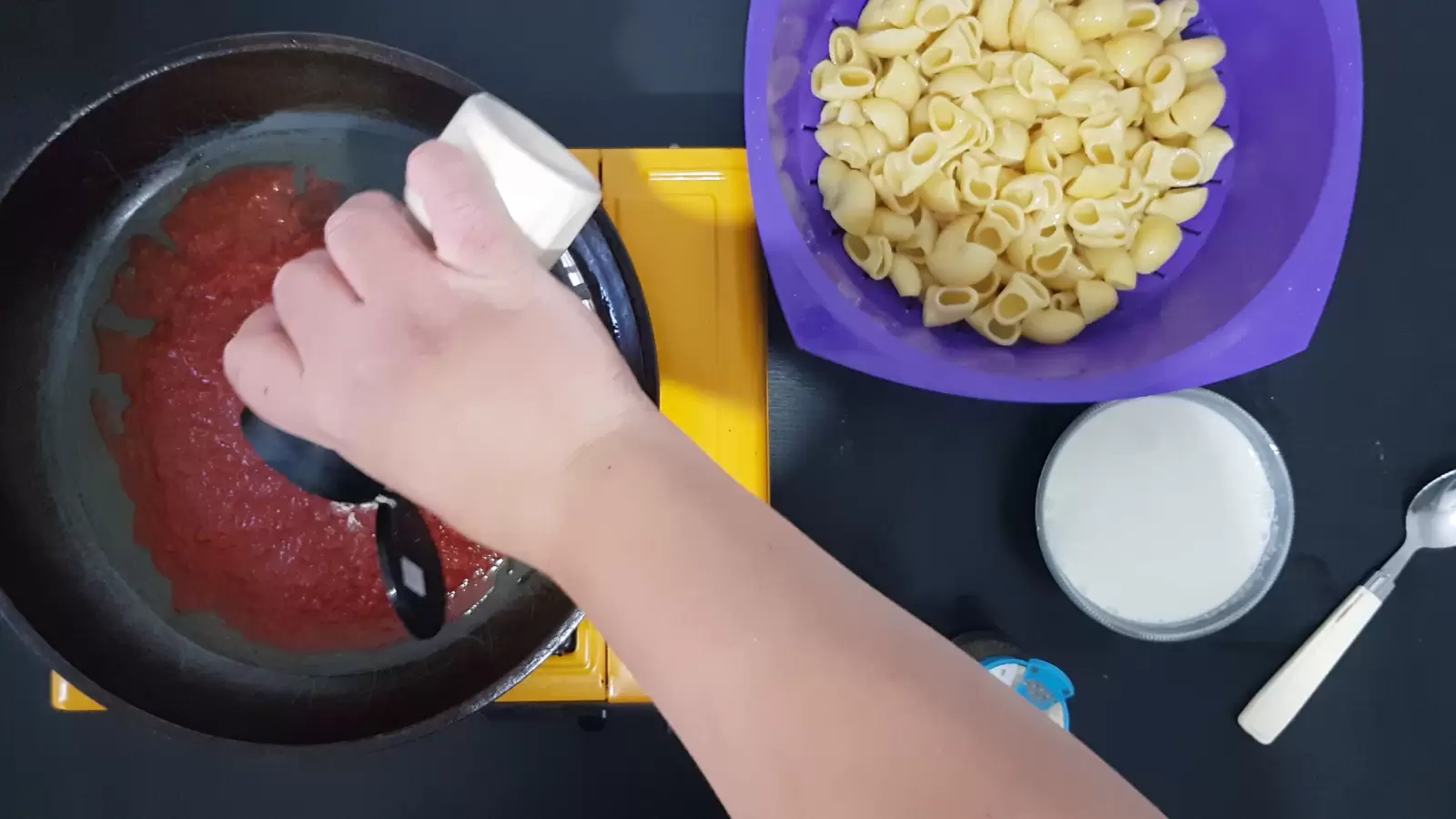 A bowl with pasta, a pan with crushed tomatoes, a jar of minced garlic, a bowl with whipped cream, a spoon