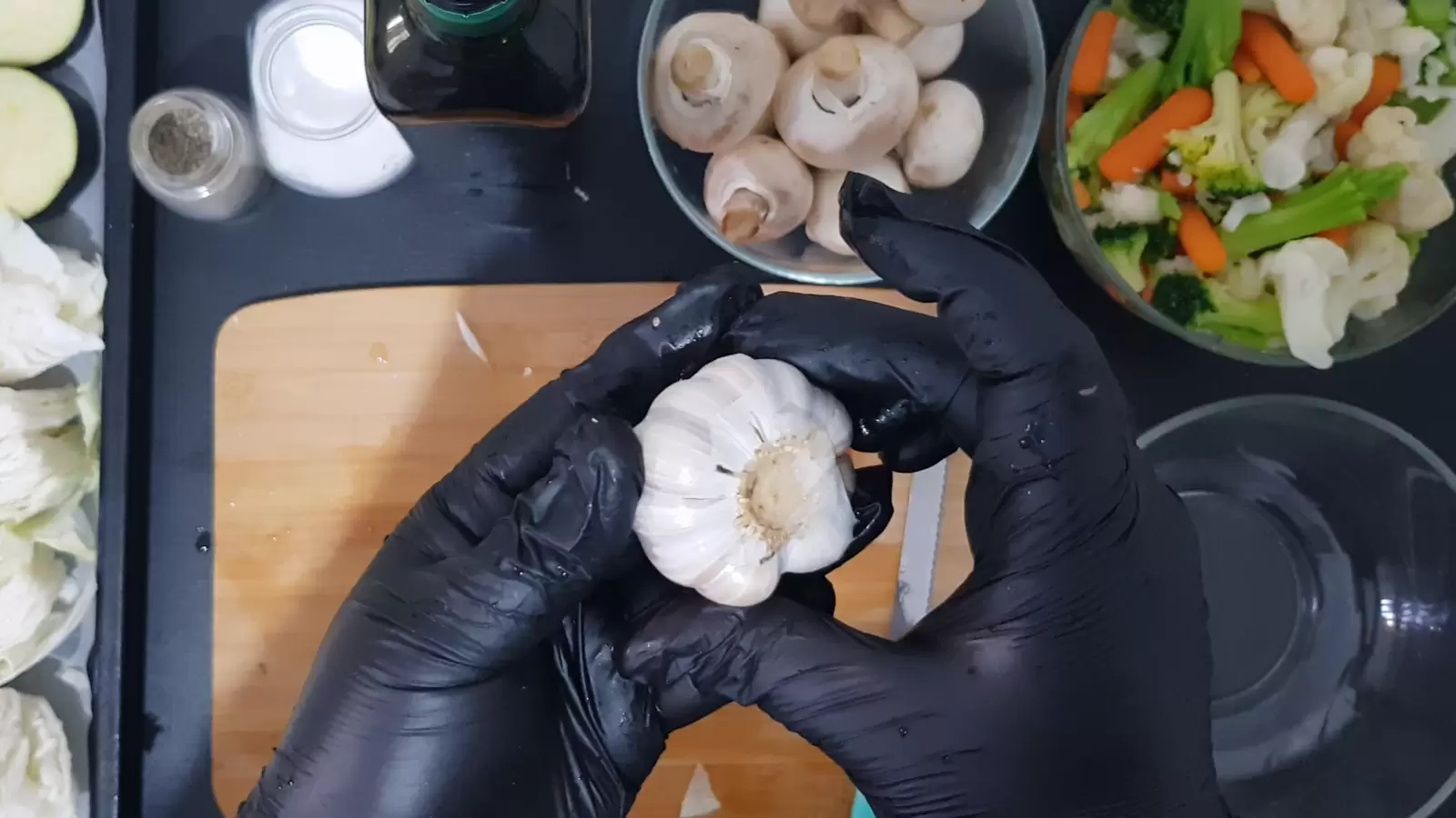 A bowl with cauliflower, carrots and broccoli, a bowl with mushrooms, a bottle of olive oil, a jar of salt, a jar of pepper, an empty bowl. Chopping board, garlic