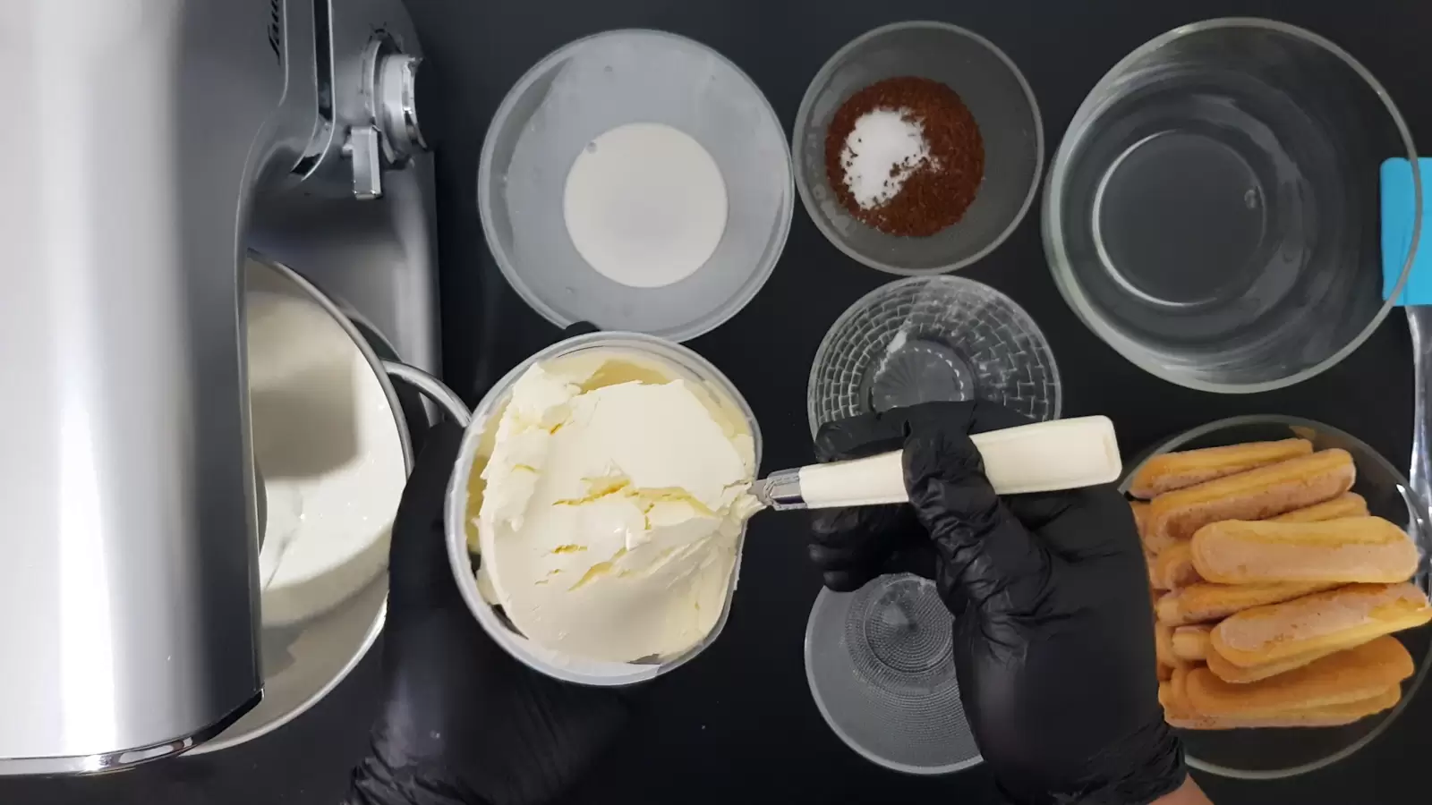 A bowl of water, a bowl of coffee and sugar, a bowl of ladyfingers, a mixer bowl with cream and a whipping balloon, a box of mascarpone and a spoon