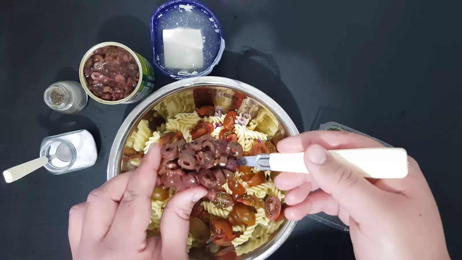 A bowl with pasta and tomatoes, a box with Bulgarian cheese, a can with olives, a spoon with olives, a jar of salt, a jar of pepper