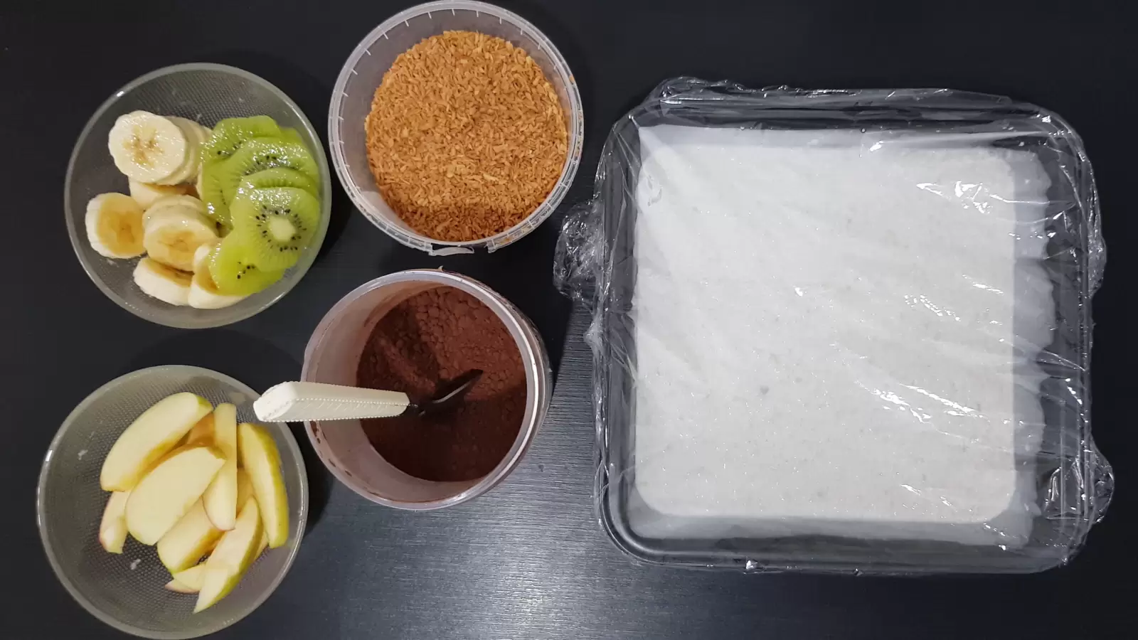 Bowl with kiwi and banana, bowl with sliced ​​apples, box with coconut chips, box with cocoa powder, mold with oatmeal