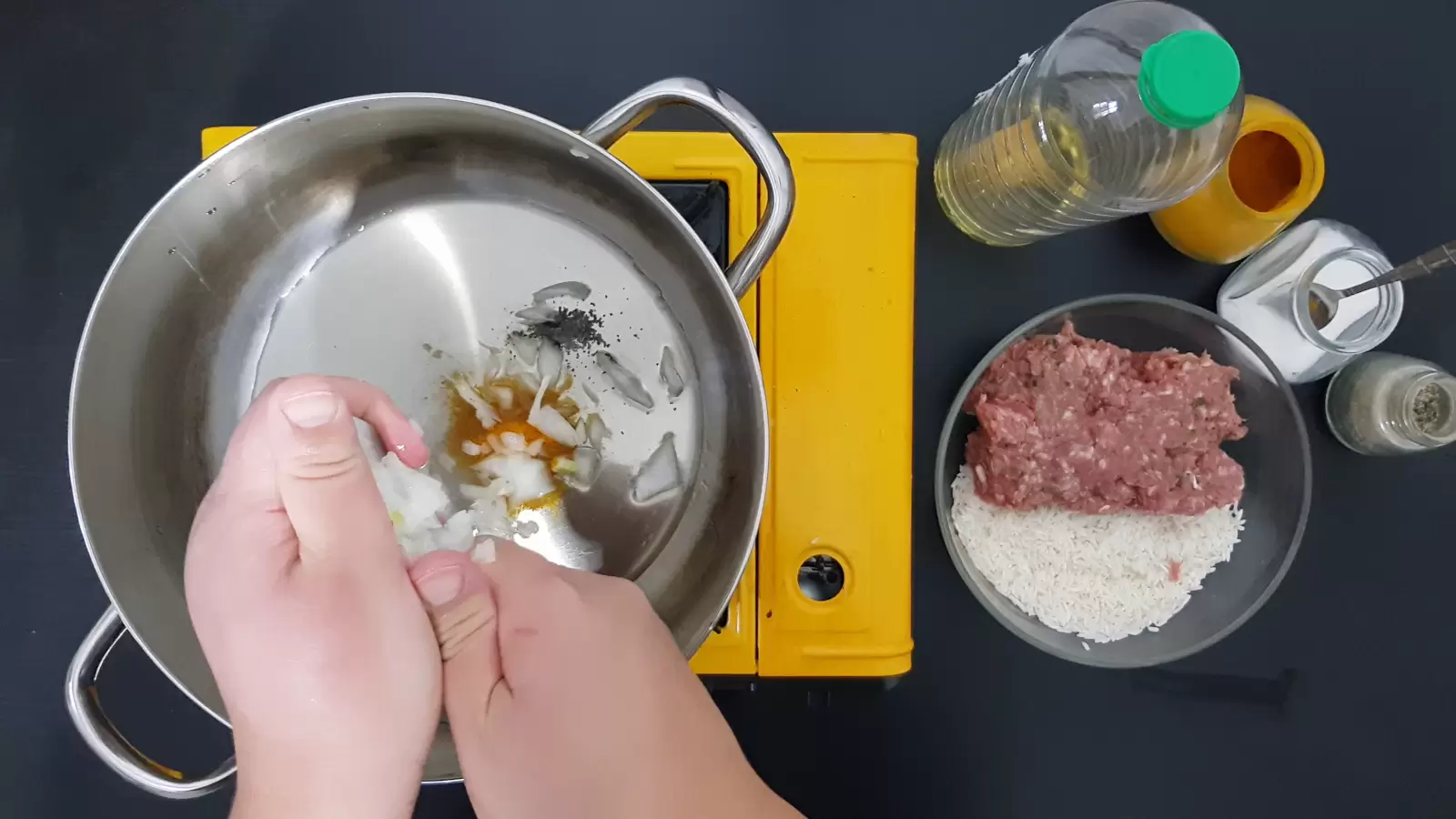 Jar with turmeric, jar with salt, jar with black pepper, half an onion, bowl with minced meat and rice, oil, pot