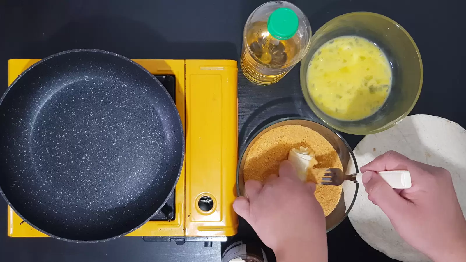 Chocolate-filled tortilla in bowl with breadcrumbs, bowl with egg, bottle of oil, stove and frying pan
