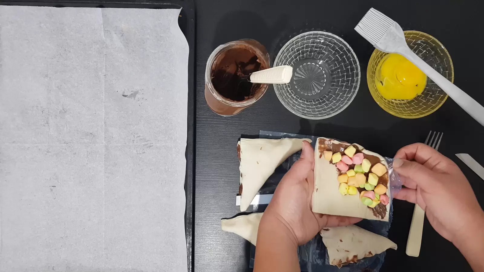 A bowl with an egg and a brush for spreading, an empty bowl, chocolate spread, a baking sheet with baking paper, puff pastry divided into squares with chocolate spread on each end, a fork