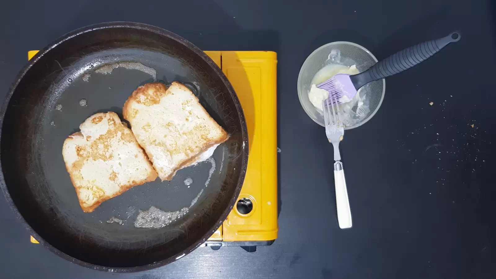 2 slices of bread in frying pan, bowl with butter and brush, fork