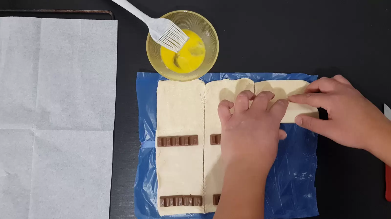 Six Kinder Bueno, rolled puff pastry, a bowl with an egg and a brush, baking paper in an oven tray