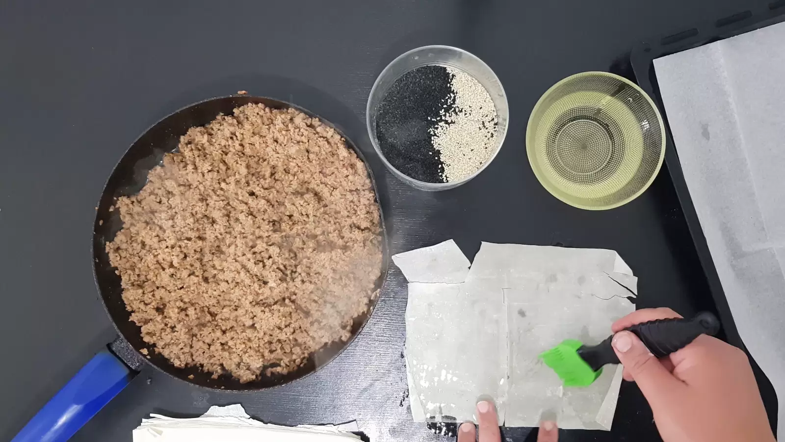 Bowl with oil, bowl with white and black sesame, pan with beef, sheet of phyllo, brush