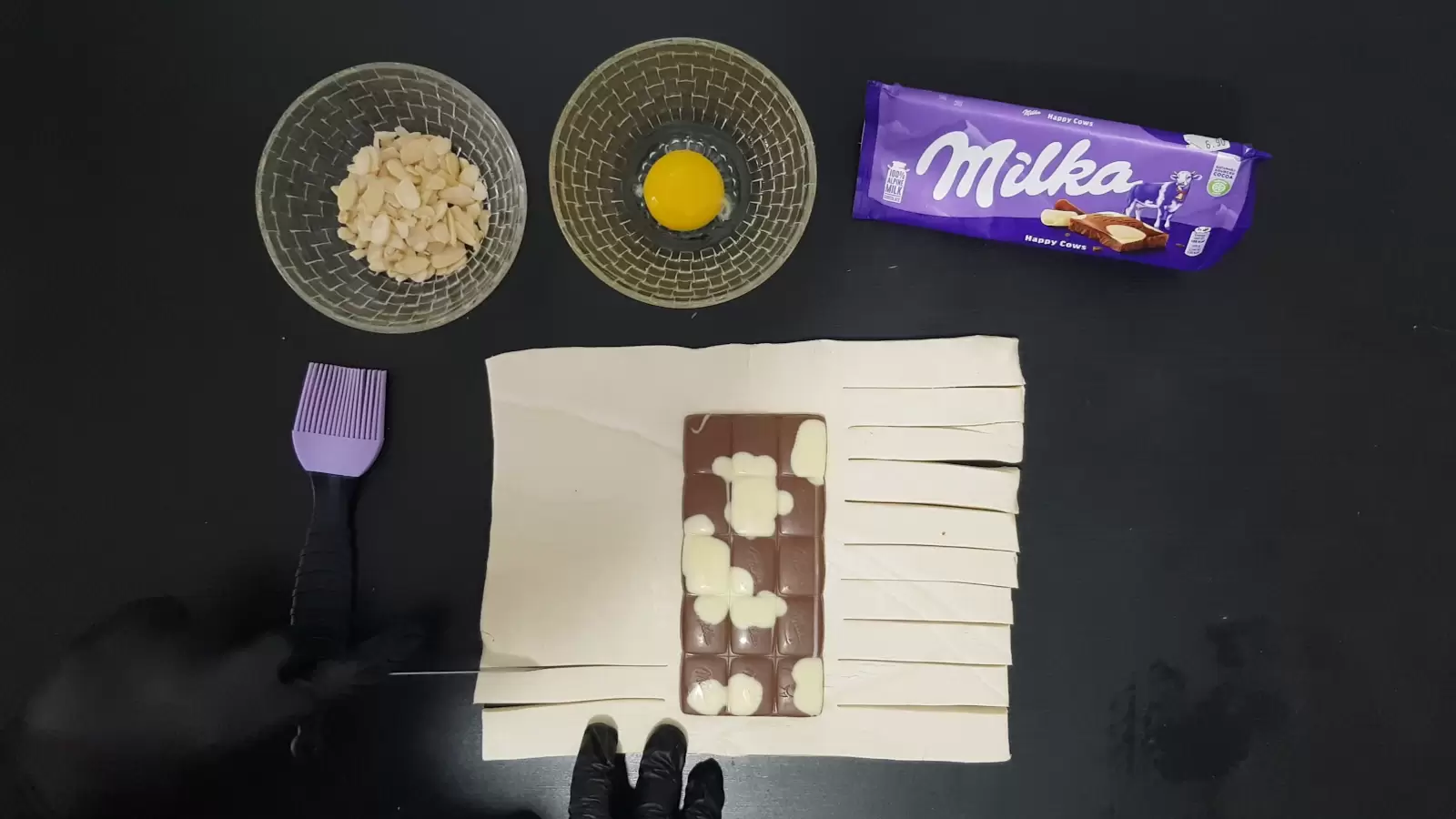 pack of Milka chocolate, bowl with egg, bowl with almonds, puff pastry with Milka chocolate, knife that cuts into strips on the left side, brush for spreading