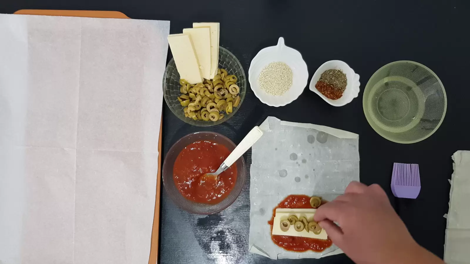 Bowl with oil, bowl with spices, bowl with sesame seeds, bowl with olives and yellow cheese, Bowl with tomato sauce, filo dough with tomato sauce, cheese, olives and herbs, brush, spoon and baking sheet with baking paper