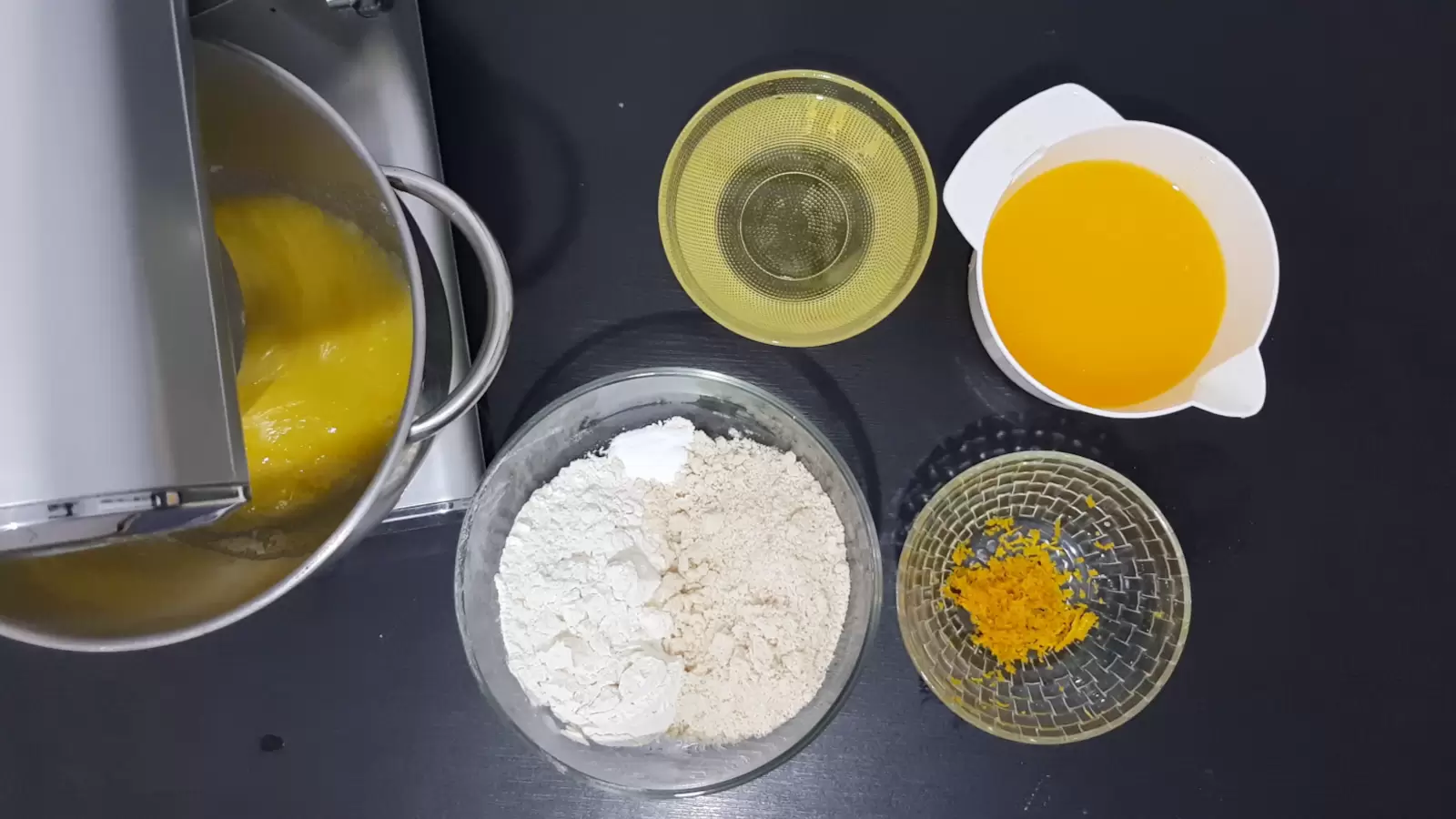 Mixer bowl with mixture, bowl with orange juice, bowl with oil, bowl with flour and almonds, bowl with orange zest