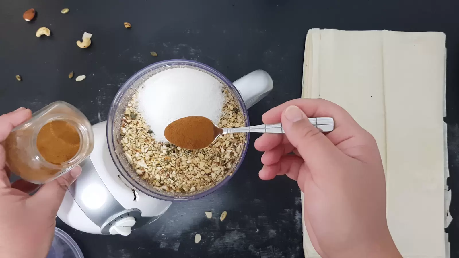 Filo sheets, a blender with ground nuts and sugar, a spoon and a jar with cinnamon