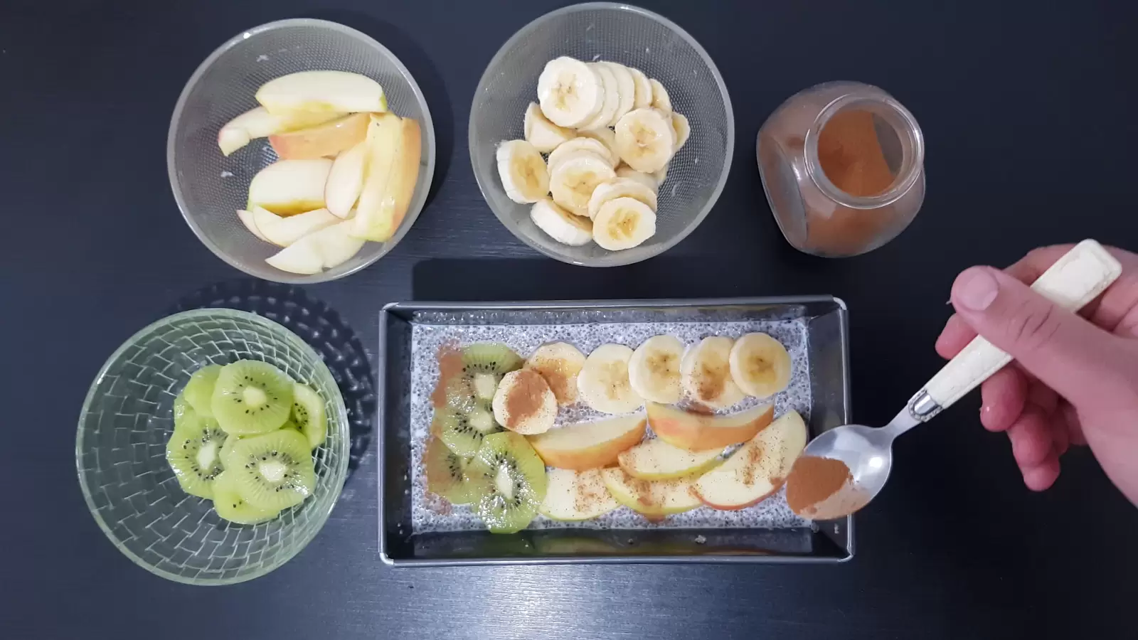 Cinnamon jar, cinnamon spoon, bowl with bananas, bowl with apples, bowl with kiwi, mold with chia pudding and fruit