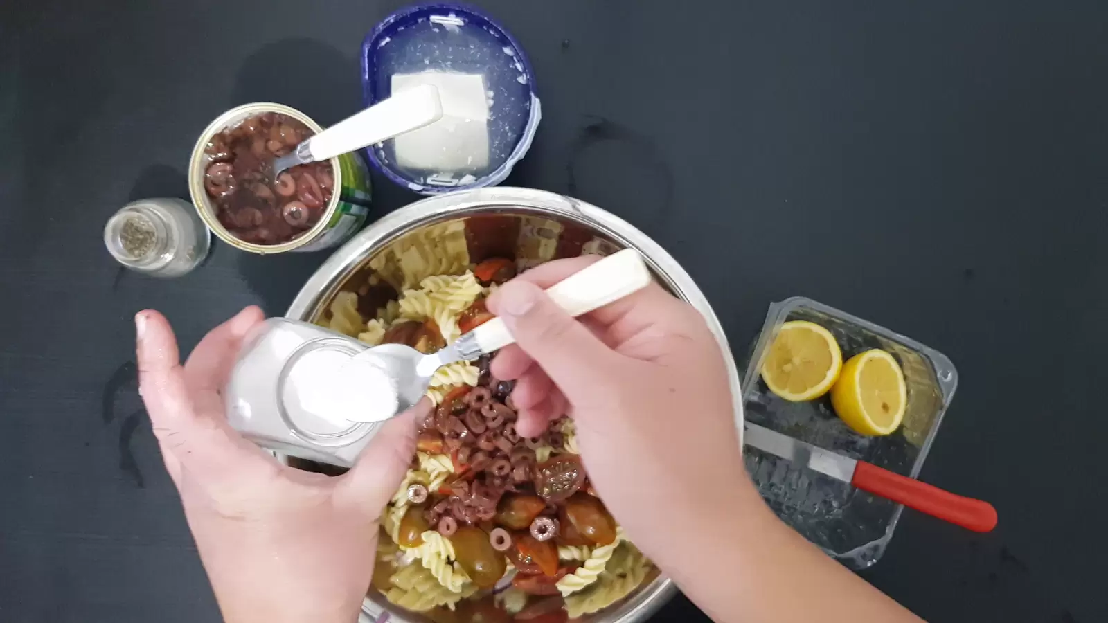 A bowl with pasta and olives, a box with Bulgarian cheese, a can with olives, a spoon in a jar of salt, a jar of pepper, a box with a lemon and a knife