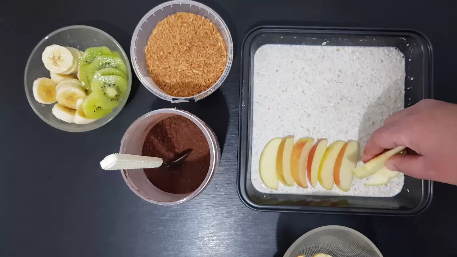 A bowl with kiwi and banana, a box with coconut chips, a box with cocoa powder, a mold with oatmeal and apple slices