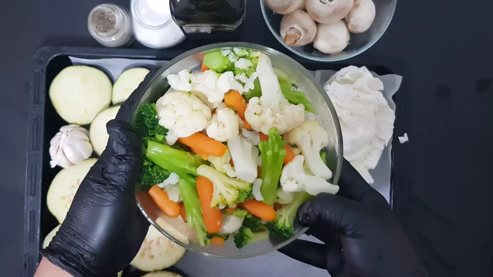 A bowl with cauliflower, carrots and broccoli, a bowl with mushrooms, a bottle of olive oil, a jar of salt, a jar of pepper.