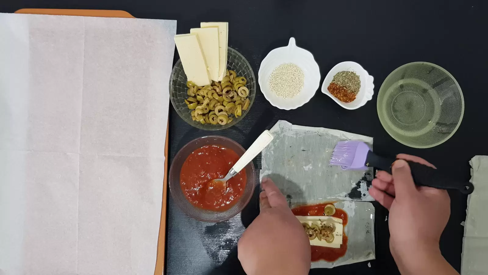 Bowl with oil, bowl with spices, bowl with sesame seeds, bowl with olives and yellow cheese, bowl with tomato sauce, filo dough with tomato sauce, cheese, olives and herbs, brush spreads oil on filo dough, spoon and baking sheet with baking paper