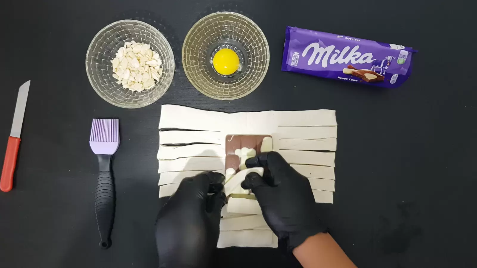 Pack of Milka chocolate, bowl with egg, bowl with almonds, puff pastry with Milka chocolate, with strips on the right and left side, brush for application, knife